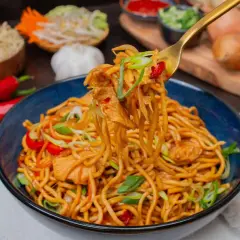Bami Goreng mit Hühnchen, garniert mit Frühlingszwiebeln und Chili. Daneben sind Zutaten wie roter Chili, Knoblauch und Zwiebeln zu sehen.