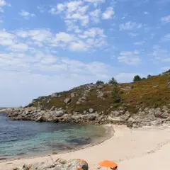 Playa rocosa y arena dorada en **Cabo Udra**, con aguas cristalinas y un cielo azul con nubes.