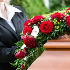 Czerwone róże, symbol ostatniego pożegnania, zdobią drewnianą trumnę. Osoba w czarnym garniturze trzyma bukiet.