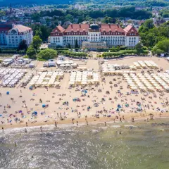 Widok z lotu ptaka na zatłoczoną plażę z białymi parasolami i hotelami w tle.