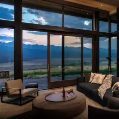 Cozy living room in a modern mountain house, featuring a fireplace, plush seating, and expansive windows with a breathtaking mountain vista at dusk.