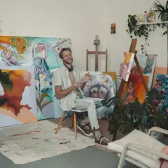 A smiling artist sits in his vibrant artist studio, surrounded by colorful canvases and plants.