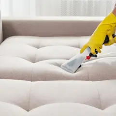 Gelbe Handschuhe führen ein Reinigungsgerät über ein helles Sofa. Tiefenreinigung für ein makelloses Sofa.
