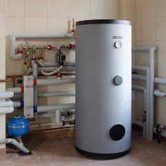 Duży, szary bojler z rurami i zaworami w pomieszczeniu technicznym.