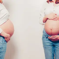 Dwa zaokrąglone brzuchy w ciąży, obie kobiety w białych koszulach i dżinsach, delikatnie dotykają swoich brzuchów.
