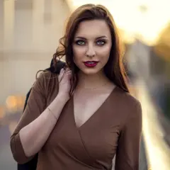 Portret młodej kobiety z burzą brązowych włosów, w brązowej sukience, z kurtką na ramieniu. Ta fotografia ludzi uchwyciła jej spojrzenie.