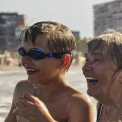 Chłopiec w goglach i starsza kobieta śmieją się na plaży. Wakacje bez zmartwień dzięki najlepszemu ubezpieczeniu turystycznemu.