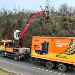 Ein orangefarbener LKW mit einem roten anbaukran schneidet Äste von einem Baum. Daneben steht ein Anhänger mit der Aufschrift "Der Biomasse Profi".