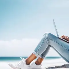 Junge Frau mit Laptop am Strand, genießt die Sonne und das Meer. Ein perfekter Ort für ihr IU Erasmus-Projekt.