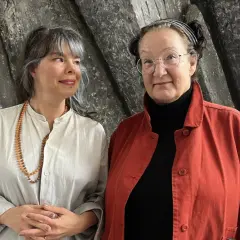 Två kvinnor står framför en grå, strukturerad vägg. Den ena, med grått hår och en ljus skjorta, bär en rad pärlor. Den andra, med glasögon och en röd jacka, ler mot kameran. Kanske är det Monika Fagerholm som samtalar med en vän.