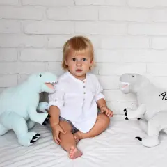 Małe dziecko siedzi między dwoma pluszowymi dinozaurami. Jasnoniebieski dinozaur po lewej i szary dinozaur z imieniem "Antoś" po prawej. Te przytulanki dla dzieci wyglądają na miękkie i zapraszające.