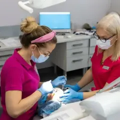 Dwie dentystki w maseczkach pracują przy pacjencie. Jedna z nich podaje narzędzia, druga wykonuje zabieg, stosując lidokainę znieczulenie miejscowe.