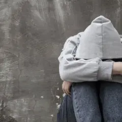 Osoba w kapturze siedzi przy szarej ścianie, przytulając kolana. W jej życiu pojawiła się **kokaina w moczu**, symbolizująca trudne wybory i ich konsekwencje.