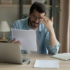 Zmartwiony mężczyzna z laptopem i dokumentami. Czyta umowę, która może dotyczyć pożyczki na wysoki procent.