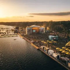 Zachód słońca nad jeziorem, gdzie odbywa się festiwal. Tłumy ludzi bawią się przy scenie, a nad wodą cumują łodzie. To miejsce, gdzie rodzi się sława rap stacja.
