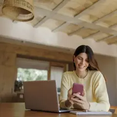 Uśmiechnięta kobieta korzysta z telefonu, sprawdzając mbank księgowanie przelewów. Obok leży laptop.
