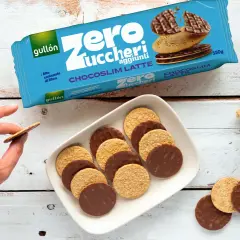 Biscotti senza zucchero Gullón Chocoslim Latte, alcuni ricoperti di cioccolato, su un piatto bianco. Una mano ne prende uno.