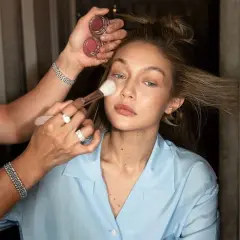 En makeupartist applicerar rouge på en kvinna som håller två små burkar med rouge. Kvinnan har sitt hår uppsatt och bär en ljusblå skjorta.