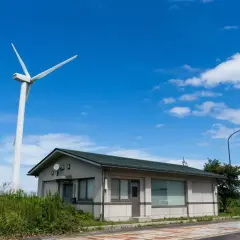 Wiatrak na tle błękitnego nieba z chmurami. Obok niewielki budynek. Zastanawiasz się, ile kosztuje postawienie turbiny wiatrowej?