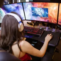 Frau mit Kopfhörern spielt an drei Monitoren. Ein "pile of shame" an Aufgaben wartet, doch jetzt zählt nur das Spiel.