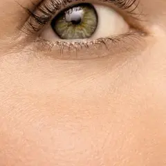 Nahaufnahme eines Auges mit grüner Iris und langen Wimpern. Die Haut wirkt blass, fast wie eine fahle Gesichtsfarbe, und zeigt feine Poren.