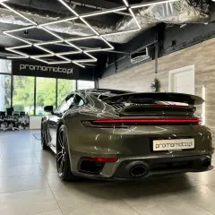 Ciemnozielone Porsche 911 w garażu z nowoczesnym oświetleniem i stojącym obok pionowym banerem.