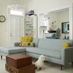Un salotto accogliente con divano azzurro, cuscini decorati e un cane bianco. L'ambiente è dominato da **colori pastello** e dettagli vintage.