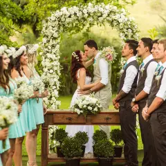 Novios besándose bajo un arco de flores, rodeados de damas de honor y padrinos.