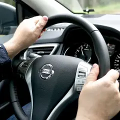 Manos al volante de un Nissan, sintiendo la vibración que indica porque vibra el volante del coche al conducir por un camino rural.