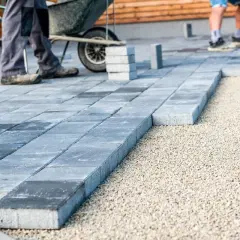 Układanie kostki brukowej na podbudowie z kruszywa. Widać pracowników i taczkę, przygotowanie podłoża to klucz do trwałości, jaki beton pod kostkę jest tu niepotrzebny.