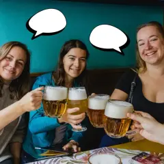 Trzy uśmiechnięte kobiety wznoszą kufle piwa. Jedna z nich mówi "Dzień dobry po czesku!".