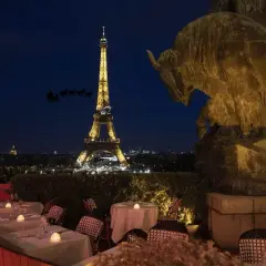 Vue nocturne de Paris avec la Tour Eiffel illuminée. Un bar ouvert le 25 décembre, avec des tables dressées et une statue de taureau. Le traîneau du Père Noël traverse le ciel.
