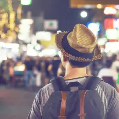 Mężczyzna w kapeluszu i z plecakiem idzie przez nocny targ, otoczony rozmazanymi światłami.