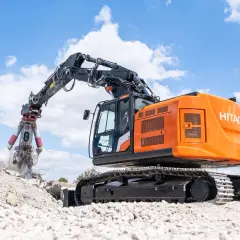 Pomarańczowa koparka do rozbiórek Hitachi z chwytakiem pracuje na gruzowisku pod błękitnym niebem z chmurami.