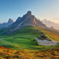 A winding road leads to a mountain lodge, perfect for hiking the Dolomites, Italy. Lush green hills meet jagged peaks under a soft sunrise.