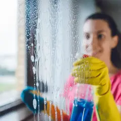 Frau mit gelben Handschuhen sprüht Reinigungsmittel auf ein Fenster, um es zu putzen.