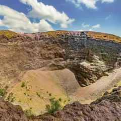 Il cratere del Vesuvio, con le sue pareti stratificate e il cono di detriti al centro, sotto un cielo azzurro con nuvole bianche.