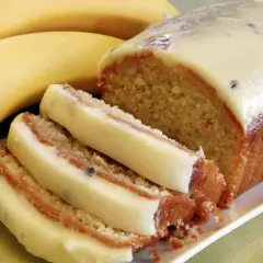 Wilgotne ciasto bananowe z kremową polewą i kawałkami owoców, obok dojrzałych bananów.