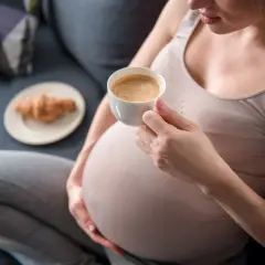 Kobieta w ciąży delektuje się kawą rozpuszczalną, siedząc na kanapie z rogalikiem na talerzyku obok.