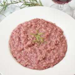 Aksamitne risotto z czerwonym winem, udekorowane gałązką rozmarynu, prezentuje się apetycznie na białym talerzu. Obok widoczny widelec i kieliszek wina.