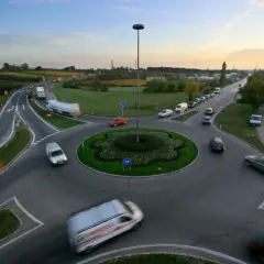 Ruchliwe rondo z wieloma rodzajami rond, samochodami i ciężarówkami. W tle widać pola i niebo o zachodzie słońca.