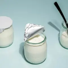 Tre burkar med hemmagjord yoghurt på ljusblå bakgrund. En är helt försluten, en annan är delvis öppnad och en tredje har en sked i sig.