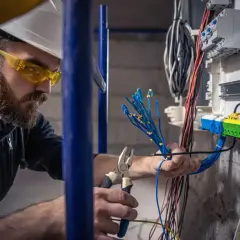 Elektryk w okularach ochronnych i kasku pracuje przy skrzynce z przewodami. Sprawdza, co daje sep g1, podłączając niebieskie kable.