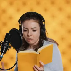 Eine Synchronsprecherin mit Kopfhörern liest ein Buch vor Mikrofon. Sie fragt sich, wie hoch wohl ihr Synchronsprecher Gehalt sein wird.