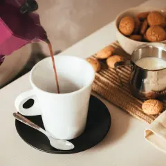 Un buon caffè fumante viene versato da una moka viola in una tazza bianca, accompagnato da biscotti e latte.