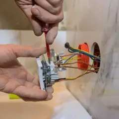 Ręce majsterkowicza montują gniazdko, przykręcając śrubki. W ścianie widać kolorowe przewody elektryczne, gotowe do podłączenia.