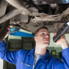 Mechanik w niebieskim kombinezonie bada podwozie auta latarką, przygotowując się do wymiany sworznia wahacza.