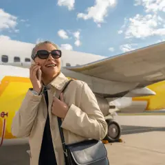 Uśmiechnięta kobieta rozmawia przez telefon przy samolocie. Ciekawe, czy w samolocie jest internet, by móc sprawdzić pocztę.
