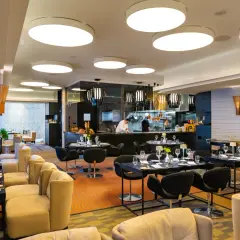 Elegancka brasserie z widokiem na kuchnię, gdzie szef kuchni przygotowuje dania. Stoliki nakryte do kolacji, wygodne fotele i nowoczesne oświetlenie tworzą przytulną atmosferę.