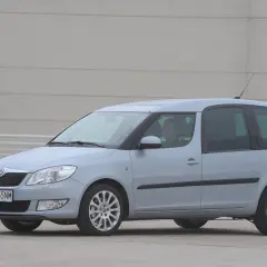 Jasnoszary Skoda Roomster z kierowcą za kierownicą stoi na betonowym placu przed szarym budynkiem.